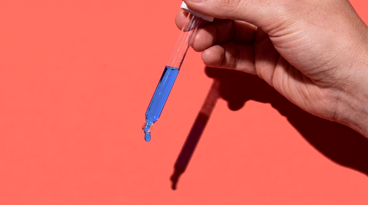A hand holding a glass pipette of blue GHK-Cu serum against a coral background